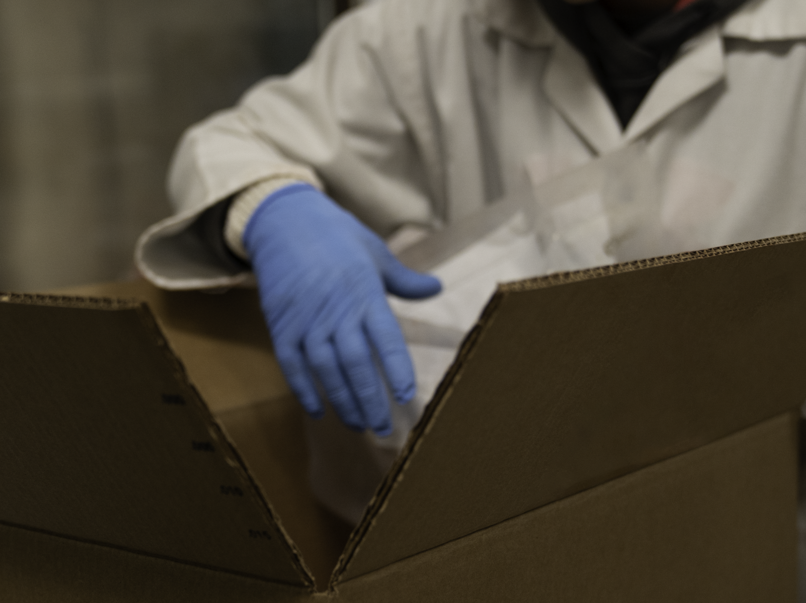 Person wearing white lab coat and blue gloves pack items into a cardboard box.