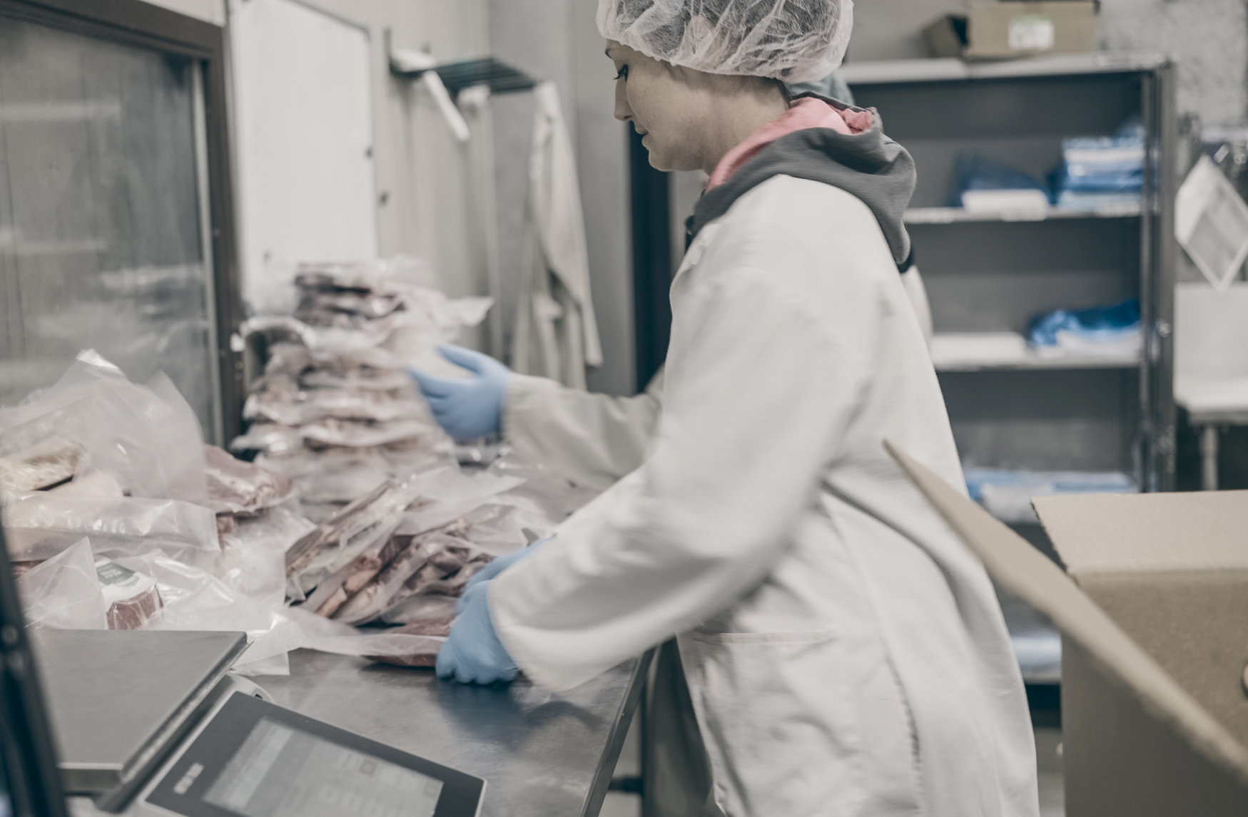 Woman in protective gear and lab coat works to package meat at a livestock processing facility.