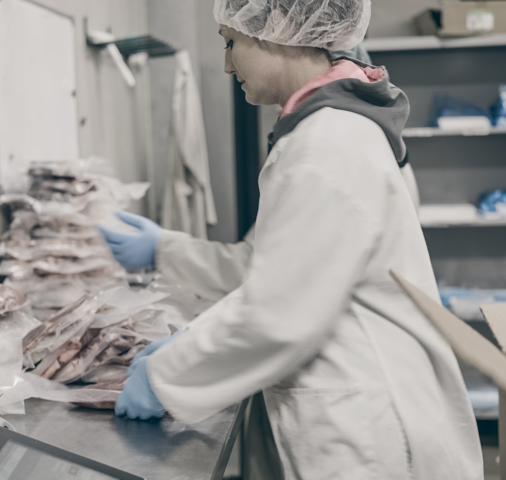 Woman in protective gear and lab coat works to package meat at a livestock processing facility.